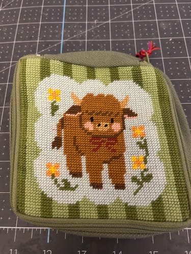 Customer's completed Highland Cow needlepoint pouch featuring a stitched brown cow with orange flowers on a green background.