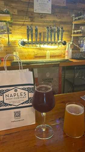 Customer's Naples Soap Company shopping bag from the Soap Sampler Set placed on a dark wooden bar next to two glasses.