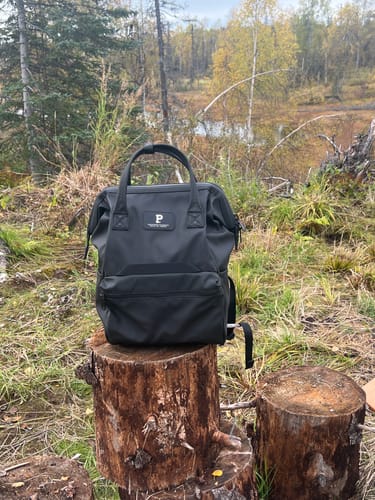 Customer's black Cascade Backpack resting on a tree stump in a forest clearing.