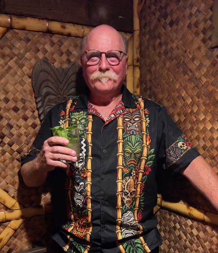 Reviewer wearing the Monsters & Mai Tais Vintage Button Up shirt while holding a cocktail in a tiki-themed bar.