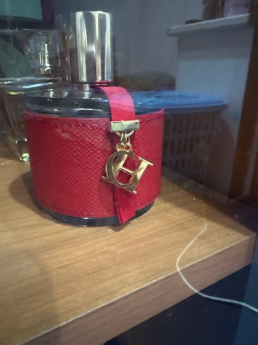 Customer's Carolina Herrera perfume bottle with a red textured leather wrap and gold CH logo charm on a table.