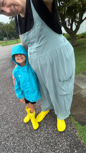 Reviewer and child wearing bright yellow Brisk I barefoot rain boots while standing on a paved outdoor walkway.