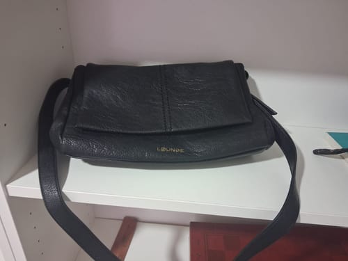 Customer's Cartera Bandolera Diseño Costuras, una cartera negra con solapa y correa, colocada sobre un estante blanco.