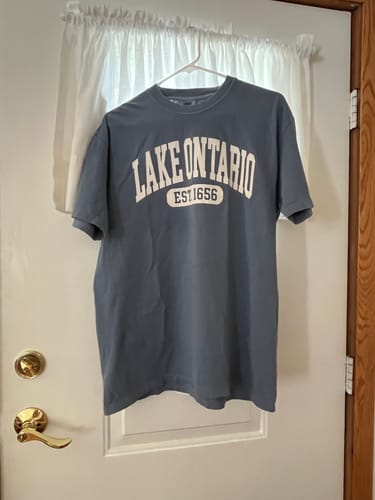 Reviewer's light denim blue Lake Ontario varsity T-shirt hanging on a hanger against a white door.