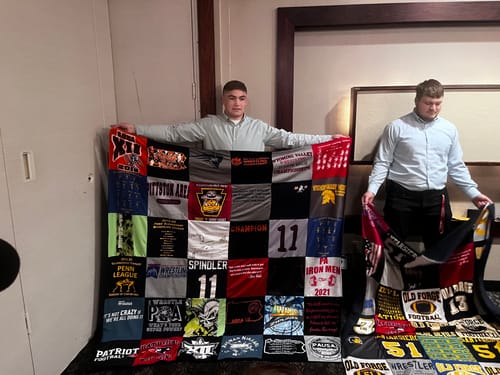 Customer's Project Repat T-Shirt Quilt held by two people, featuring a collection of various sports t-shirts.