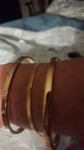 Customer's wrist wearing three gold-tone bracelets, including the Vina Bracelet, in a bedroom.