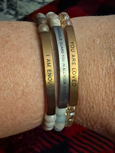 Customer's wrist wearing three beaded I AM ENOUGH bracelets with engraved metal bars reading I AM ENOUGH, ANGELS GUARD YOU, and YOU ARE LOVED.