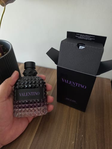 Customer's Valentino Uomo Born in Roma perfume bottle held by hand, next to its open black box on a wooden surface.