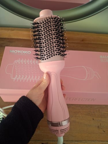 Customer's hand holding a pink Moyoko Magnitude 2-in-1 Hot Air Brush in front of its product packaging box.