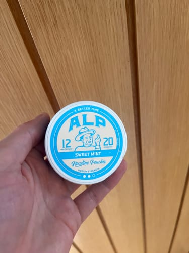 Reviewer's hand holding a blue and white ALP Sweet Mint nicotine pouch tin against a wooden wall background.
