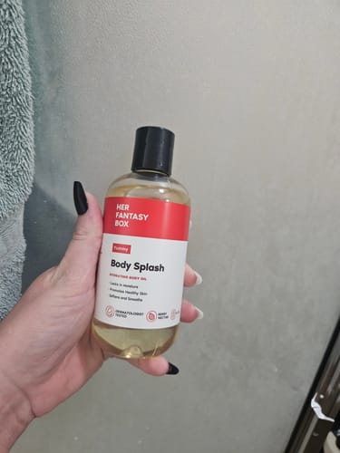 Reviewer's hand holding a clear, round bottle of Yummy Body Splash oil against a grey wall.