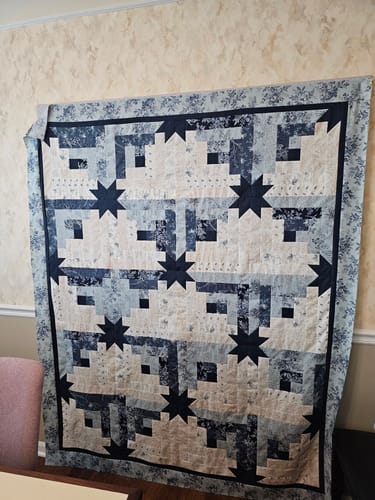 Customer's Maggie's Cabin quilt featuring blue and cream star and log cabin patterns hanging on a wall.