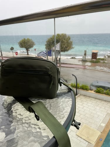 Customer's green Crossbody Organizer resting on a glass table with a rainy coastal view in the background.