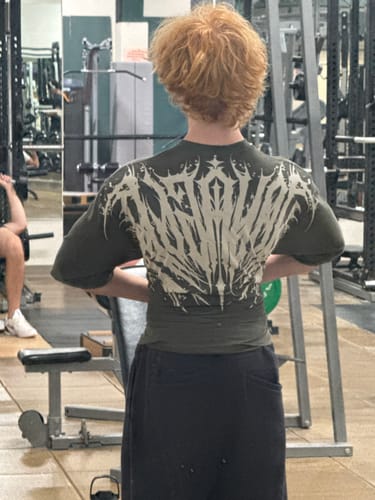 Customer's dark green Chaos Tee with tattoo-inspired back graphic, worn in a gym.