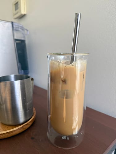 Customer's FLUR iced coffee glass filled with coffee and a metal straw, sitting on a wooden surface.