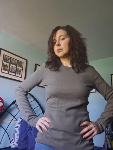 Reviewer wearing a mushroom brown Italian cashmere crew neck jumper with hands on hips, standing in a bedroom.