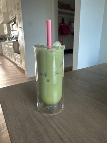 Customer's iced matcha in a clear FLUR glass with a pink straw on a wooden table.
