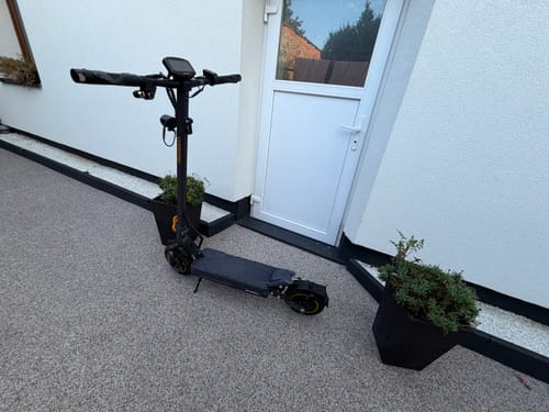 Reviewer's black Ausom K20 Pro e-scooter parked on a gravel patio beside a white house door.
