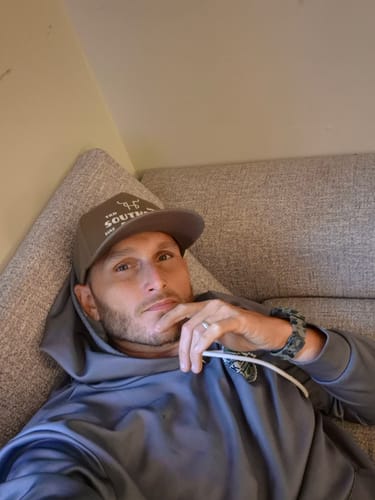 Reviewer wearing a brown Hat of the Month Crew baseball cap while relaxing on a sofa.