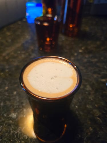 Customer's amber FLUR Cortado Glass filled with coffee topped with foam, sitting on a dark counter.