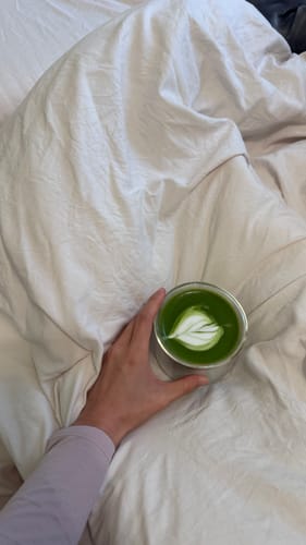 Customer's hand holding a clear FLUR Latte Glass filled with a green matcha latte featuring leaf foam art on a white bed.