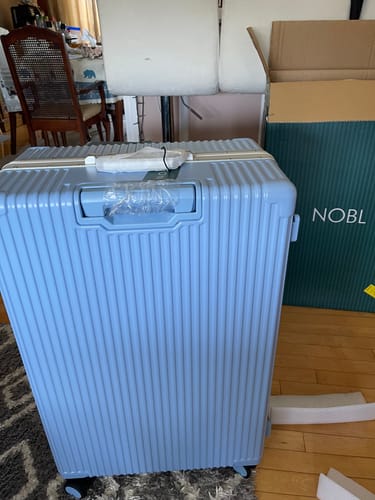 Customer's new light blue Check-In All-In-One suitcase standing in a room next to its packaging box.