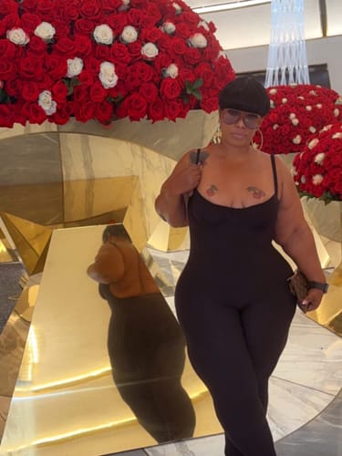 Customer's Naked Open Back Jumpsuit in black worn in front of large red floral arrangements.