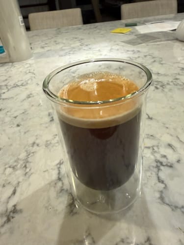 Customer's clear FLUR Tasting Glass filled with dark, frothy coffee on a marble surface.
