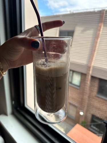 Customer's hand holding a clear, double-walled FLUR espresso glass filled with iced coffee and a metal straw.