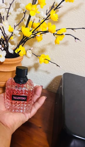 Customer's hand holding a clear, textured glass bottle of Valentino perfume in front of yellow flowers.