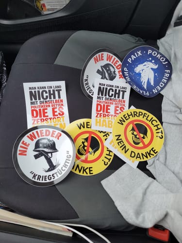 Customer's Nie wieder Kriegstüchtig Aufkleber stickers in black, white, red, and yellow, sitting on a dark car seat.