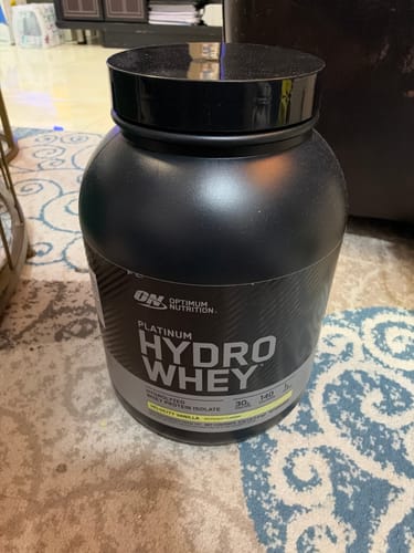 صورة لمراجع لعلبة مسحوق بروتين Optimum Nutrition Platinum Hydro Whey ذات اللون الأسود موضوعة على سجادة منقوشة.
