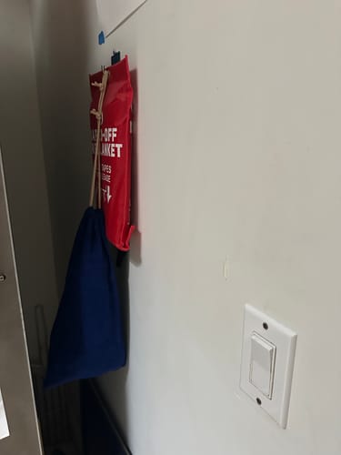 Customer's red fire blanket bag and blue drawstring bag hanging on a white wall near a light switch.