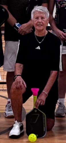 Reviewer wearing white CUURT Muuv court shoes while kneeling on a wooden court holding a pickleball paddle and ball.
