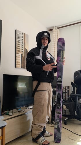 Customer's khaki baggy snow pants worn with a black jacket and a ski, standing in a room.