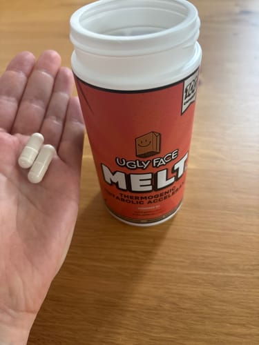 Reviewer's hand holding two white capsules next to a red UGLYFACE MELT supplement bottle.