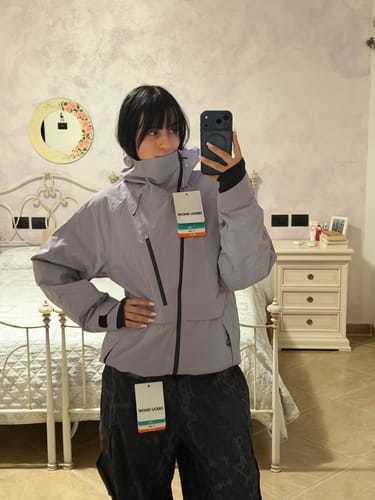 Reviewer wearing a lavender waterproof snow jacket with tag, in a bedroom setting.