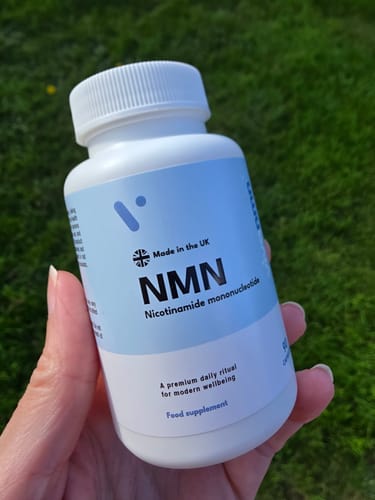 Main de la cliente tenant un flacon blanc de gélules Pure NMN 500 mg sur fond d'herbe verte.