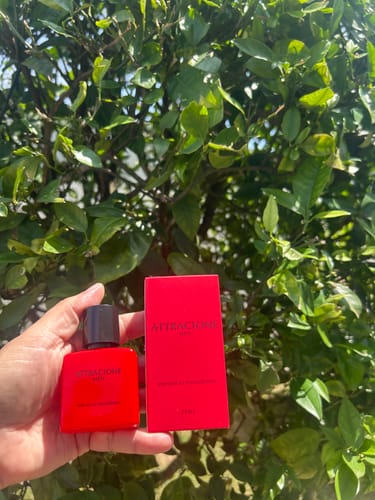 Customer's Attracione Men perfume bottle and red box in a hand, with green leaves in the background.