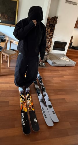 Reviewer standing in a room, wearing black baggy snow pants with skis on the floor nearby.