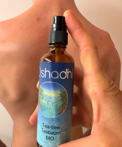 Reviewer's hand holding an Oshadhi Teebaum-Hydrolat bottle in front of a person's back.