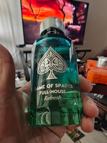 Reviewer's hand holding a green glass Jo Milano Full House Refresh perfume bottle with a spade emblem