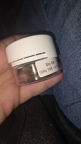 Customer's hand holding a clear, lidded jar containing THC-A 50% Hash Dry Sift.