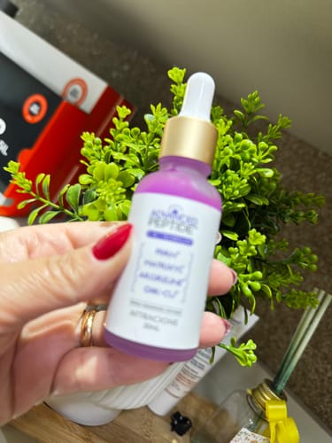 Customer's hand holding a purple Advanced Peptide serum bottle with a gold dropper cap against green foliage.