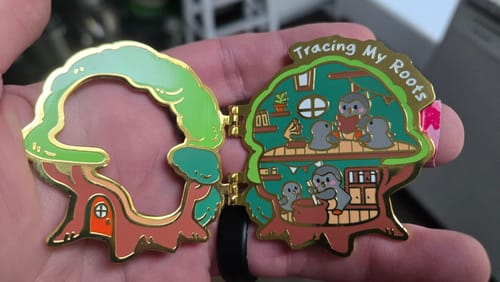 Customer's Tracing My Roots pin opened in hand, showing a tree-shaped enamel design with gold detailing.