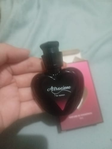 Customer's hand holding the dark, heart-shaped Attracione Sensual perfume bottle in front of its product box.