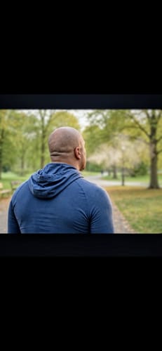 Reviewer in a blue hoodie exhaling a faint cloud of vapor outdoors in a park setting.