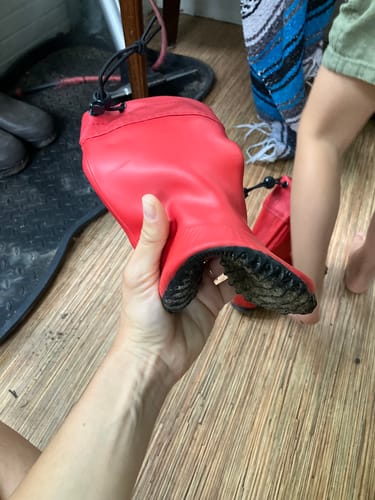 Customer's hand holding a flexible, red Kid's Brisk I barefoot rain boot, showing its pliable, textured sole.
