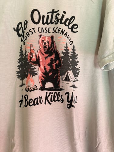 Customer's white Go Outside T-shirt featuring a graphic of a bear with a bottle by a campfire and a tent in the forest.