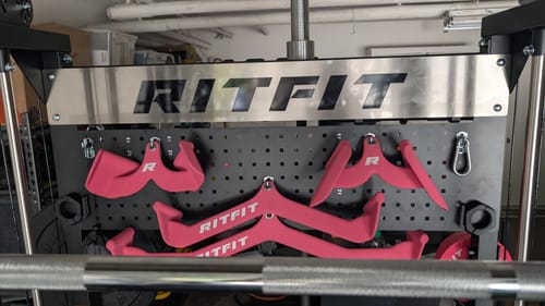 Reviewer's RitFit PBM1 Pegboard Attachment mounted on a gym rack, holding pink gym equipment handles.
